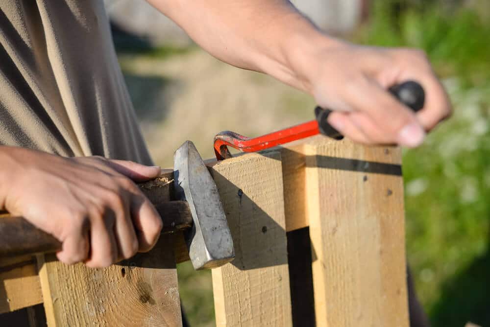 Hands using a hammer and pry bar to fix a wood fence picket