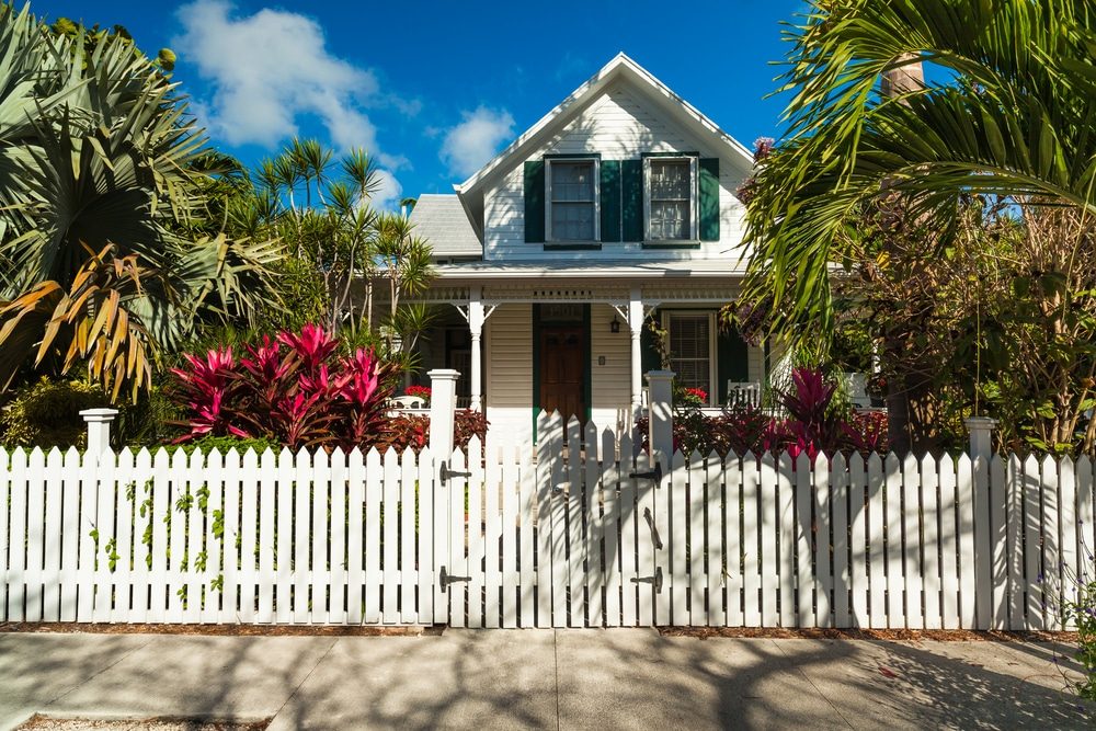 A white cottage with green shutters behind a white picket fence and gate, with palm trees and red tropical plants