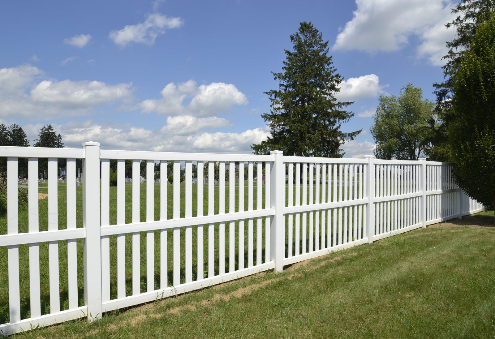 White vinyl picket-style fence running along a green lawn under a blue sky
