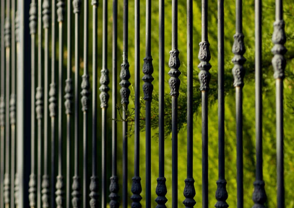 Black metal fence pickets with carved decorative collars in front of green foliage