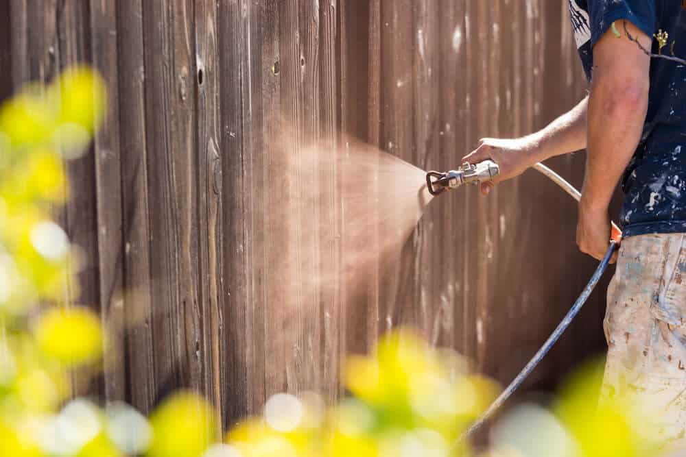 A worker spraying stain onto a brown wood fence with a paint sprayer