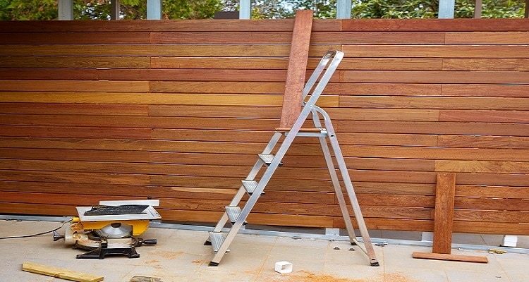 A tall horizontal wood fence with a ladder, a miter saw, and loose boards in front of it