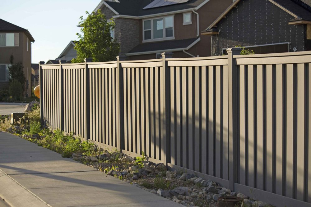 A gray-brown vinyl fence with narrow gaps between boards running along a sidewalk in front of two houses