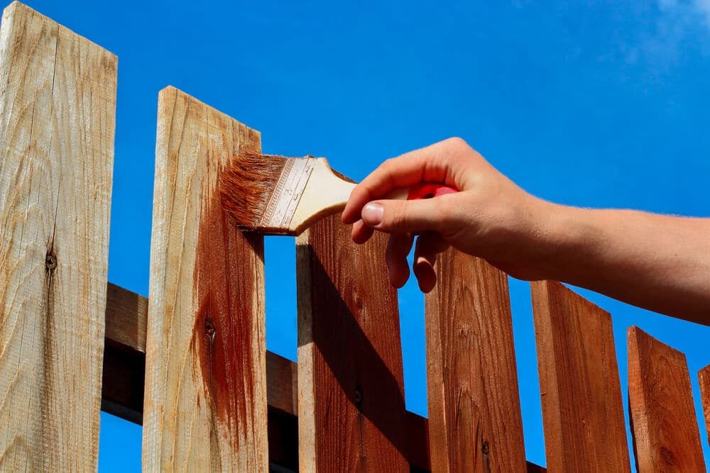 A hand brushing reddish stain onto tall wood fence pickets against a blue sky