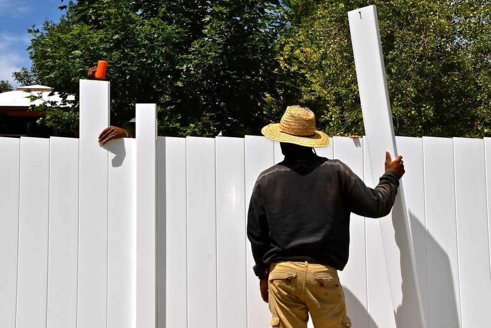 A worker holding a white vinyl fence panel against a white vinyl privacy fence