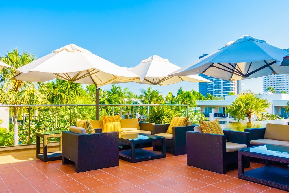Large cream and blue umbrellas shading wicker sofas and tables on a sunny terrace with palm trees