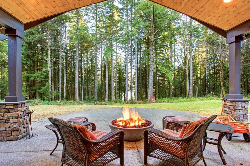 A covered patio with a wood ceiling, stone-base columns, and wicker chairs around a fire bowl