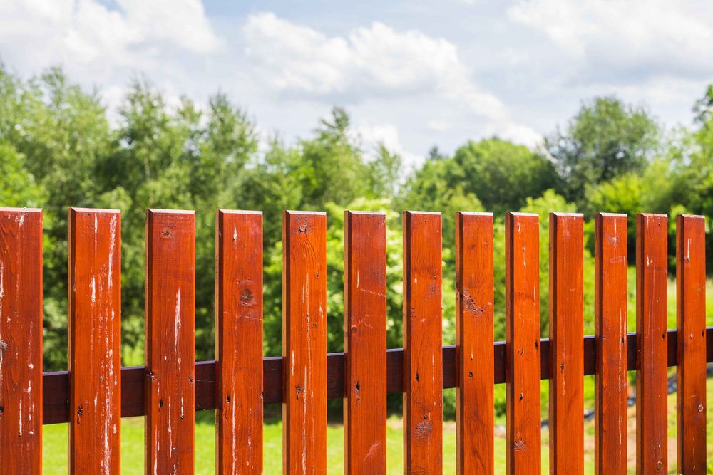 A wood picket fence finished in a deep red-brown stain with green trees and blue sky behind it