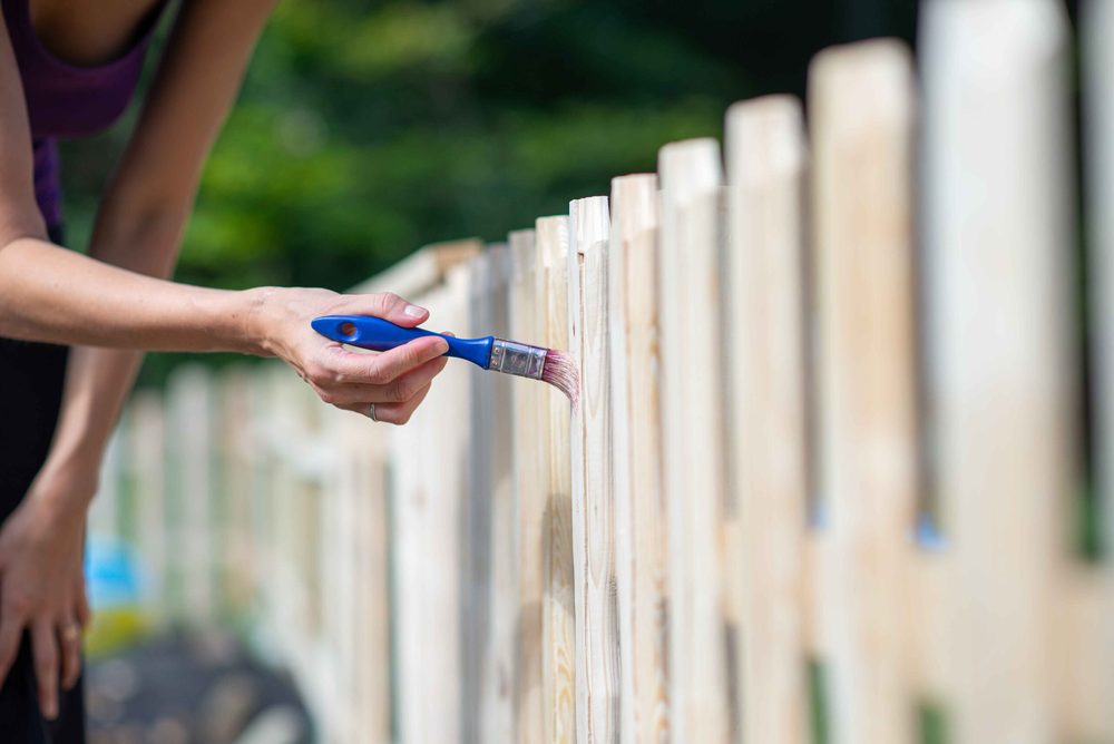 A person brushing clear sealer onto a raw wood picket fence with a blue-handled brush