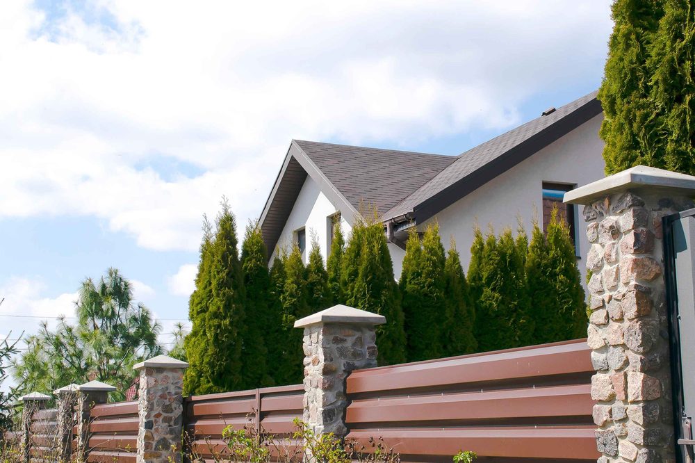 A brown metal fence with stone columns in front of a white house and tall green evergreen shrubs