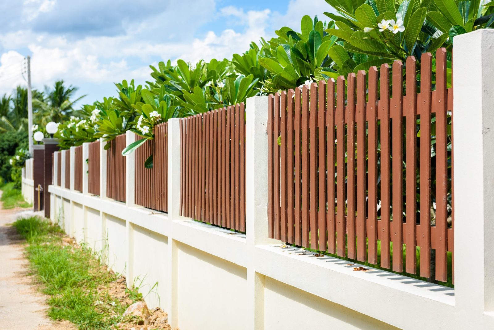 Brown wooden picket fence set on top of a cream stucco wall with tropical plants behind it