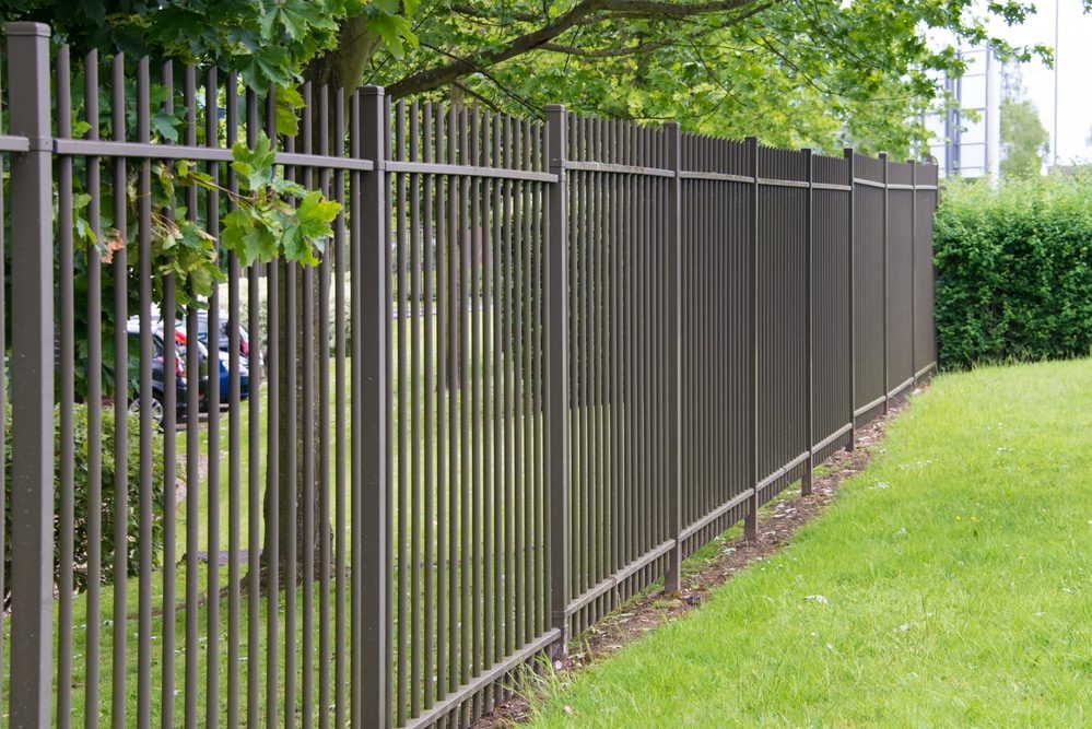 Dark bronze metal fence with vertical pickets running along a green lawn and trees